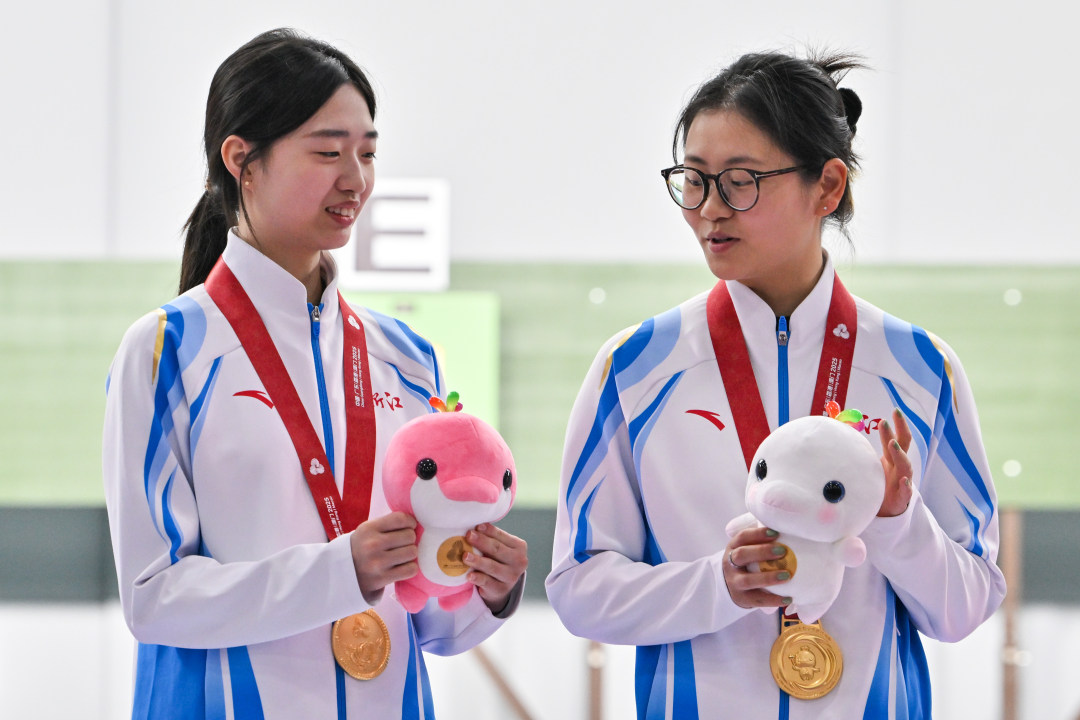 2025年10月14日,十五运会女子10米气步枪团体决赛颁奖仪式上,浙江队队员黄雨婷(左)与队友韩佳予(右)比划十五运会吉祥物。